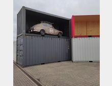 Ein altes Auto mit der Nummer "62" steht auf einem grauen Frachtcontainer, der Teil einer gestapelten Anordnung aus weiteren Containern in unterschiedlichen Farben (grau, rot) ist. Der Bereich ist ein gepflasterter Außenplatz unter wolkigem Himmel.