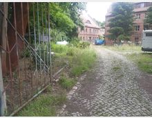 Eingezäuntes Gelände mit Kopfsteinpflasterweg, der zu mehreren alten, roten Backsteingebäuden führt. Im Hintergrund stehen zwei Gebäude mit Satteldächern, umgeben von Bäumen, und ein geparkter blauer Transporter sowie ein Wohnmobil rechts im Bild.