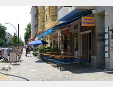 Straßenszene mit einer Reihe von Geschäften und Restaurants, darunter ein indisches Restaurant mit einem blauen Schild und Außensitzplätzen mit Holzbänken. Entlang des Gehwegs stehen Fahrräder, ein blauer Sonnenschirm und Blumenkübel, während Fußgänger und Radfahrer die Straße nutzen; mehrstöckige Wohnhäuser im Hintergrund.