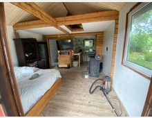 Holzvertäfelter Innenraum eines kleinen Hauses mit Schrägdach. Im Vordergrund ein Bett mit weißer Bettwäsche, ein Schrank und ein Staubsauger; im Hintergrund ein Fernseher, ein Tisch mit Dekor und ein Ofen auf einer Glasplatte. Außen erkennt man durch das Fenster einen Garten mit Bäumen.