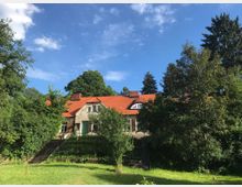 Ein einstöckiges Haus mit roten Ziegeldächern und einer hellen Fassade, umgeben von dichtem Grün und hohen Bäumen. Im Vordergrund befindet sich ein gepflegter Garten mit Rasen und schmalen Treppen, die zum Gebäude führen.