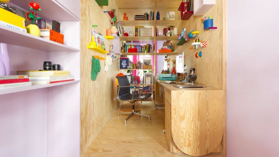 Narrow built-in study nook finished in light plywood, with a small desk, shelves, and wall-mounted cubbies filled with books and colorful objects. A black swivel chair sits in the center, and the space is integrated into a room with pale pink shelving on the left.