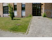 Backsteingebäude mit großen Fenstern und moderner Eingangstür aus Glas und Metall. Davor befindet sich eine kleine Grünfläche mit einem jungen Baum und ein gepflasterter Weg, der zum Eingang führt.