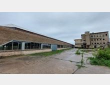 Ein verlassener Industriekomplex mit einer langen Halle aus Ziegelmauerwerk und großen Fenstern links sowie einem mehrstöckigen, verwitterten Gebäude rechts. Der Bereich dazwischen ist eine betonierte Freifläche mit vereinzeltem Grasbewuchs.