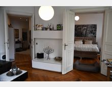 Innenansicht einer Wohnung mit Holzdielenboden. Zentraler Blick auf eine geöffnete Tür, die in ein Schlafzimmer mit Doppelbett und großformatigem Stadtbild an der Wand führt. Im Vordergrund ein weißes Regal mit Dekorationsgegenständen und ein silberner Kerzenhalter, daneben ein Hundebett auf dem Boden.