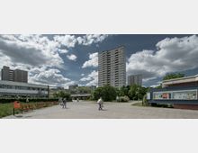 Wohngebiet mit Hochhäusern im Hintergrund, einer Grünfläche mit Bäumen und Sträuchern in der Mitte sowie einem Weg, auf dem Menschen laufen. Im Vordergrund sind rote Bänke, eine Informationstafel und ein niedrigeres Gebäude sichtbar.