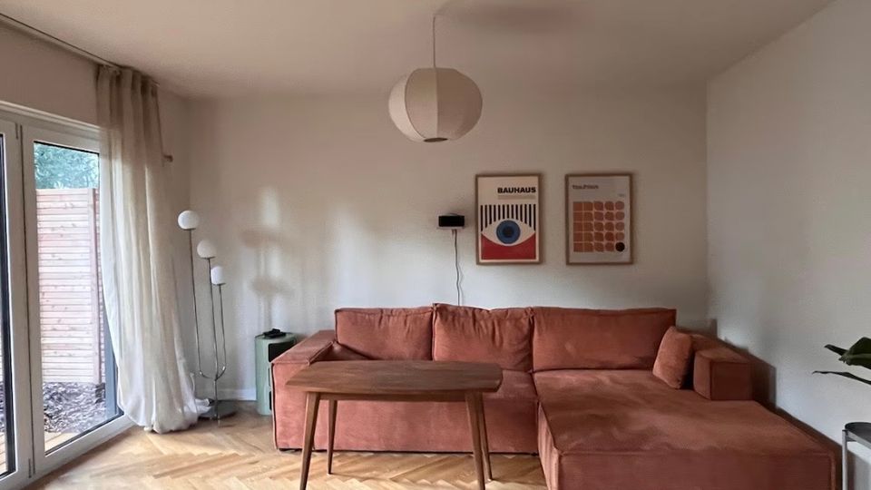 Wohnzimmer mit einem Ecksofa in Terrakottafarbe, einem kleinen Holztisch und einem hölzernen Fischgrätenparkettboden. An der Wand hängen zwei gerahmte Poster, daneben steht eine Stehlampe mit mehreren kugelförmigen Lampenschirmen. Links führen bodentiefe Fenster mit weißen Vorhängen auf eine Terrasse.