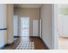 Heller Flur in einer Altbauwohnung mit hohen Decken und Stuck, beige gestrichenen Wänden, weißen Türrahmen und blau abgesetzten Sockelleisten. Dunkler Holzboden mit einem Abschnitt aus gemusterten Fliesen; mehrere Türen führen in weitere Räume, am Ende ist ein Raum mit großen Fenstern sichtbar.