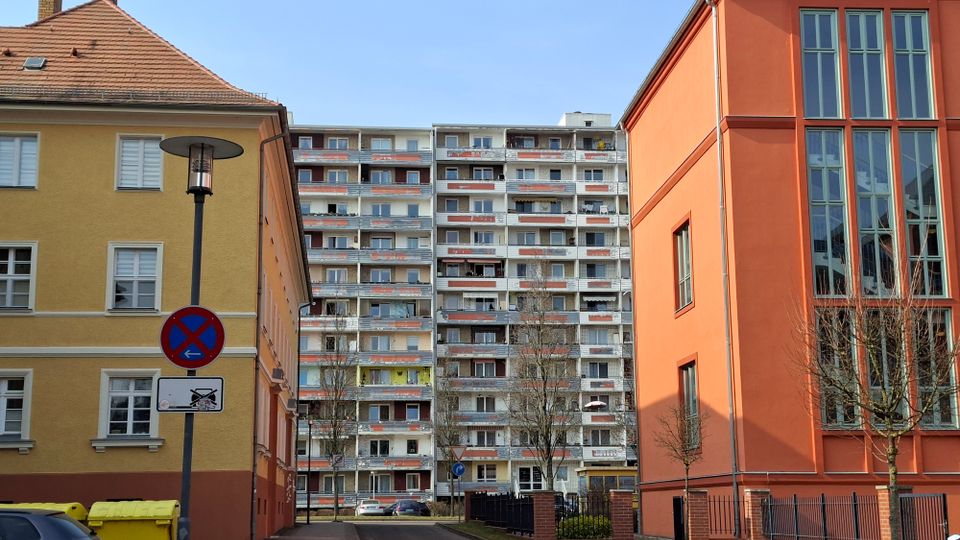 Blick in eine schmale Wohnstraße, eingerahmt von einem gelben Altbau links und einem orangefarbenen Gebäude mit hohen Fensterflächen rechts. Im Hintergrund steht ein mehrstöckiger Plattenbau mit vielen Balkonen; davor sind Straßenlampe, Verkehrsschild, kahle Bäume und parkende Autos zu sehen.