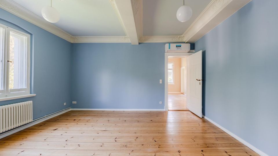 Leerer Wohnraum mit hellblauen Wänden und Holzdielenboden, links ein großes Fenster mit Heizkörper darunter. Die Decke hat dekorative Stuckelemente und zwei runde Hängelampen; rechts führt eine offene Tür in einen angrenzenden Flur.