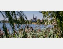 Blick durch Schilf und Bäume auf einen See mit einer Kirche im Hintergrund, deren Doppeltürme in den Himmel ragen, umgeben von Bäumen und einigen Gebäuden.