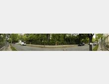 Panoramaaufnahme einer ruhigen, gepflasterten Wohnstraße, gesäumt von Bäumen und Grünflächen. Auf der linken Seite ist ein Metallzaun sichtbar, rechts stehen Autos entlang des Bürgersteigs, dahinter befindet sich dichter Baumbestand.