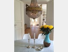 Ein elegantes Esszimmer mit einem runden, weißen Marmortisch, auf dem zwei silberne Kerzenhalter und eine Vase mit gelben Blumen stehen. Im Hintergrund ist ein Kronleuchter mit goldenen Verzierungen sowie ein Blick auf eine helle Diele mit Fliesenboden und Bücherregalen zu sehen.