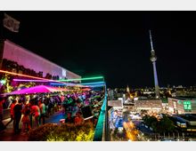 Dachterrasse mit Menschen und beleuchteten Sonnenschirmen, bunten Neonlichtern und Begrünung an den Wänden; im Hintergrund eine nächtliche Stadtansicht von Berlin mit dem beleuchteten Fernsehturm.