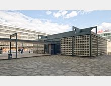 Kleines, modernes Gebäude mit einer Fassadengestaltung aus quadratischen Betonblöcken, umgeben von einer gepflasterten Fläche. Im Hintergrund sind ein mehrstöckiges Bürogebäude und einige Passanten zu sehen.