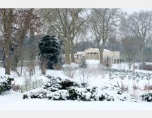 Schneebedeckter Park mit Bäumen und einem kleinen Holzsteg im Vordergrund. Im Hintergrund steht ein klassizistisches Gebäude mit Säulen, umgeben von kahlen Bäumen.
