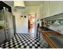 Eine helle Küche mit einem schwarz-weiß karierten Boden. An der rechten Wand befinden sich weiße Hängeschränke, eine Holzarbeitsplatte mit Gaskochfeld und eine Spüle. Links steht ein großer Edelstahlkühlschrank. Im Hintergrund ist eine offene Tür zu einem weiteren Raum mit Fliesenboden sichtbar.