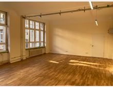 Ein leerer Raum mit hellen Wänden und einem Holzboden. Große Fenster an einer Seite lassen Tageslicht herein, daneben befinden sich Heizkörper und eine weiße Tür führt zu einem angrenzenden Bereich. Deckenleuchten und Kabelschienen sind sichtbar.