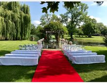 Grüne Außenfläche mit weiß dekorierten Sitzreihen und einem roten Teppich, der zu einem mit Blumen geschmückten Traubogen führt. Im Hintergrund ein Teich, Bäume und ein gepflegter Park.