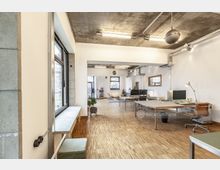 Heller Büroraum mit industriellem Design, Holzboden und Sichtbetondecke. An den Wänden befinden sich große Fenster, die den Raum mit Tageslicht füllen, und es sind moderne Arbeitsplätze mit Schreibtischen und Stühlen eingerichtet.