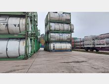 Stapel von silbernen Tankcontainern in grünen Metallrahmen auf einem Lagerplatz mit Betonboden, daneben weitere Container auf Eisenbahnwaggons. Im Hintergrund sind Bäume und gestapelte Frachtcontainer zu sehen.