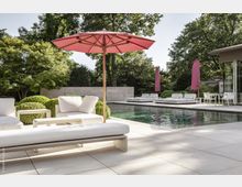 Moderner Außenbereich mit Pool, weißen Liegen, Sitzmöbeln und einem roten Sonnenschirm auf einer hellen Steinfläche, umgeben von gepflegter Bepflanzung und Bäumen. Im Hintergrund befinden sich weitere Sitzgelegenheiten sowie eine teils verglaste Gebäudefassade.