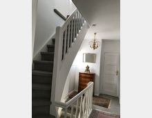 Innenraum eines Wohnhauses mit einer weißen Treppe mit Geländer, die nach oben führt. Im Hintergrund steht eine Holzkommode mit einer goldfarbenen Tischuhr, eine Wandlampe und Teppiche schmücken den Raum.