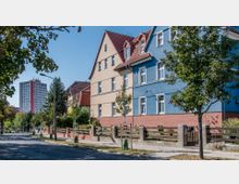 Straßenszene in einem Wohngebiet mit mehrstöckigen, farbigen Gebäuden in beige und blau sowie roten Ziegeldächern; im Hintergrund ein modernes, hohes Wohnhaus und Bäume entlang der Straße.