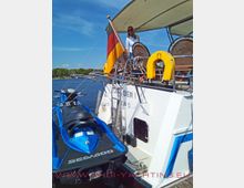 Eine motorisierte Yacht mit einem Sonnendach liegt an einem Steg, auf deren Heck ein deutscher Flaggenmast und gelbe Rettungswesten sichtbar sind. Ein blaues Jetski ist am Heck befestigt, und eine Frau steht auf dem hinteren Deck der Yacht.