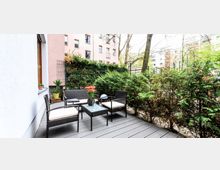 Kleine Terrasse mit Holzboden, ausgestattet mit geflochtenen Gartenmöbeln und einem Tisch mit Blumen. Im Hintergrund sind grüne Sträucher sowie umliegende Wohnhäuser sichtbar.