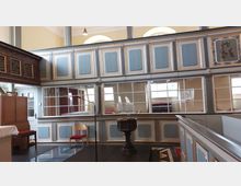 Innenraum einer historischen Kirche mit hellblauen und goldverzierten Holzelementen. Im Vordergrund steht ein Taufbecken auf einem dunklen Holzsockel, daneben ein Mikrofon und ein Holzstuhl, im Hintergrund sind Fenster und eine Empore erkennbar.