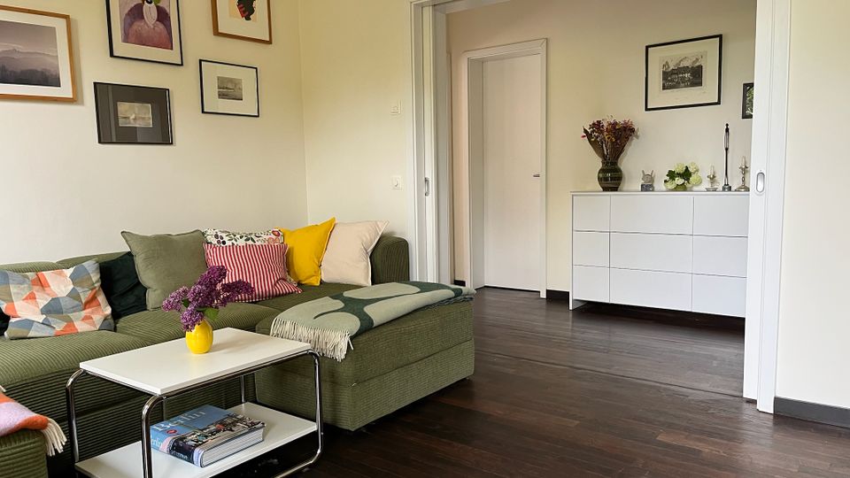 Wohnzimmer mit dunklem Holzboden, grünem Ecksofa mit bunten Kissen und kleinem weißem Beistelltisch. An der Wand hängen mehrere gerahmte Bilder; durch eine breite Schiebetüröffnung ist ein angrenzender Raum mit weißer Kommode und Dekoration zu sehen.