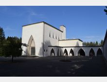 Weiße Kirche mit gotischen Elementen, einschließlich Spitzbogenfenstern und -türen. Das Gebäude steht in einem offenen Hof mit gepflasterter Fläche, einem kleinen Springbrunnen und umgeben von Bäumen.