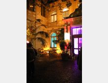 Beleuchteter Innenhof eines historischen Gebäudes mit hohen Fenstern und Bogendetails. Im Hof stehen kleine Tische und Stühle, umgeben von Pflanzen; bunte Lichtprojektionen an Wänden und Fenstern sorgen für eine stimmungsvolle Atmosphäre.