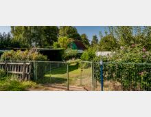 Ein Bauerngarten mit einem offenen Metallzaun im Vordergrund, umgeben von blühenden Sträuchern und großen Bäumen. Im Hintergrund steht ein kleines, grünes Haus mit einem roten Dach, eingebettet in eine ländliche Landschaft.