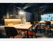 Ein modern gestalteter Café-Innenraum mit Holztischen und Stühlen, bei gedämpfter Beleuchtung. Im Hintergrund befindet sich eine beleuchtete Theke, während große Fenster Tageslicht hereinlassen und vereinzelte Blumenvasen auf den Tischen stehen.