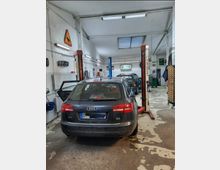 Innenraum einer Autowerkstatt mit einem dunklen Audi A6 im Vordergrund. Die Werkstatt verfügt über Hebebühnen, Werkzeuge an den Wänden und Deckenbeleuchtung; ein Ford-Logo ist im Hintergrund sichtbar.