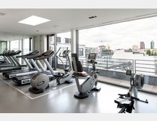 Fitnessraum mit modernen Trainingsgeräten, darunter Laufbänder, ein Fahrradergometer und ein Rudergerät, vor einer Fensterfront mit Ausblick auf eine Stadtlandschaft mit Gebäuden und Flaggenmast. Ein großer Wandspiegel verstärkt die räumliche Wirkung.
