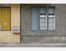 Eine Straßenfassade mit einem gelb gekachelten Bereich links, einem vergitterten Fenster und einem roten Kaugummiautomaten. Rechts ist eine graue Putzwand mit drei Fenstern, teils mit geschlossenen Rollläden, zu sehen.