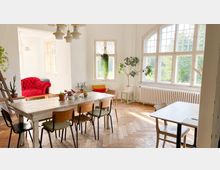 Heller, einladender Raum mit großen Sprossenfenstern, die viel Tageslicht hereinlassen, einem hölzernen Esstisch mit mehreren Stühlen und Pflanzen an den Fenstern und auf dem Tisch. Im Hintergrund ist ein rotes Sofa in einer gemütlichen Ecke zu sehen.