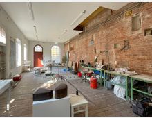 Alter Arbeitsraum mit hohen Decken, Ziegelwand und großen Fenstern, die Tageslicht hereinlassen. Im Raum befinden sich Werkbänke, Materialien und Werkzeuge sowie eine rote Holztür im hinteren Bereich.