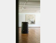 Ein minimalistischer Ausstellungsraum mit Holzboden und einer weiß gestrichenen Decke. Im Vordergrund steht eine skulpturale Installation in Form eines Baumstamms mit mehreren angebrachten Wasserhähnen, im Hintergrund hängt ein abstraktes Gemälde an der weißen Wand.