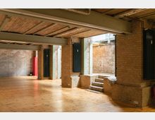 Ein Innenraum mit freiliegenden Ziegelwänden, einer Decke aus Holz und Metallträgern sowie einem Holzboden. Im Vordergrund sind schwarze Wandheizkörper, und im Hintergrund führen wenige Stufen zu einem erhöhten Bereich mit weiteren Ziegelwänden.