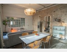 Offener Wohnbereich mit industriellem Design, gekennzeichnet durch eine unverputzte Ziegelwand, ein großes Fenster mit Glasbausteinen und eine moderne Hängelampe. Im Vordergrund steht ein Esstisch mit Holzstühlen, während im Hintergrund ein graues Sofa und ein Regal mit Geschirr zu sehen sind.