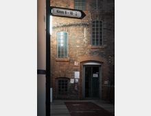 Backsteingebäude mit schmalen, rechteckigen Fenstern und einem Eingang mit Doppeltüren; darüber ein beleuchtetes Schild mit der Aufschrift „Kinos 6 - 10“. Im Vordergrund ein ebener Bodenbelag mit Markierungen, daneben ein Feuerlöscher an der Wand.