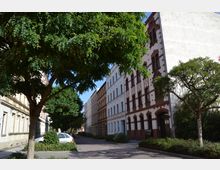 Straßenansicht mit Wohnhäusern in Altbauarchitektur, gekennzeichnet durch Verzierungen aus roten Backsteinen und helle Fassaden. Bäume säumen den Bürgersteig, und ein geparktes Auto steht am Straßenrand.