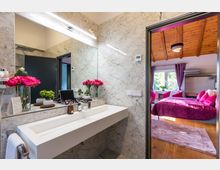 Modernes Badezimmer mit einer großen, beleuchteten Spiegelwand und weißem Waschbecken, umgeben von Marmorfliesen. Durch eine offene Tür ist ein Schlafzimmer mit Holzdecke, großen Fenstern, violetten Vorhängen und einem Bett mit magentafarbenem Bettbezug sichtbar.