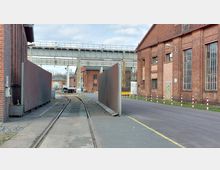 Industrielle Umgebung mit Gleisen, die zwischen roten Backsteingebäuden verlaufen. Im Hintergrund ist eine Metallbrücke zu sehen, und die Straße ist von Pollern und grünem Rasen gesäumt.