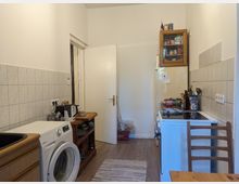 Ein schmaler Küchenraum mit weißen Wänden und Fliesen an den Wänden. Links steht eine Waschmaschine und eine Spüle, daneben ein Regal mit Geschirr. Rechts befinden sich ein Kühlschrank, ein Herd und eine Arbeitsfläche mit Küchenutensilien. Im Hintergrund ist eine geöffnete Tür zu sehen.