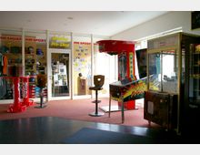 Ein Innenraum mit Automaten, darunter ein roter Boxautomat und ein Flipperautomat, sowie ein Bereich mit einer Glastür, die zu einem „Bowling Pro-Shop“ führt. Der Raum enthält außerdem Vitrinen, Verkaufsartikel, einen hohen Hocker und eine Ladentheke.